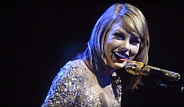 Swift seen smiling while performing on a microphone. She is wearing a bob haircut and a sparkling suit