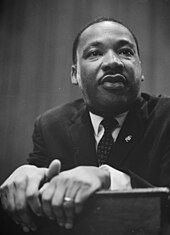 African American man in his forties, dressed in a suit and tie, leaning and grasping on the edge of a lectern