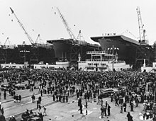 Black and white photograph of numerous people standing on a concrete surface, in front of three ships under construction with cranes on top of them