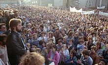 A town square is filled with thousands of people, some holding large banners, looking towards a group of people on a platform in the left foreground. A man with a beard is in the foreground in front of microphones, addressing the crowd.