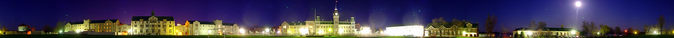 A 260-degree photo of the Royal Military College of Canada in Kingston, Ontario, on 4 May 2007. Seen is a green landscape during the night, featuring buildings made of white stone and red brick. The night sky is dark blue and purple, with the moon shining bright on the right side of the image. Photo credit: Martin St-Amant (User:S23678)