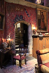 Une salle chaleureuse aux teintes rouges, avec tapisseries médiévales, tapis, fauteuil, meubles en bois et escalier de pierre en colimaçon menant à l'étage.