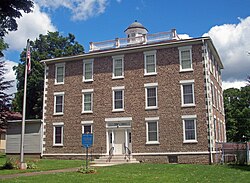 A three-story building faced in small earth-toned rocks with a rounded cupola on top. There is an American flag on a flagpole at left and a blue-and-gold historical marker in front. Above the door are the words "Town Hall".