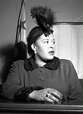 African American woman, in her late thirties, sitting in a courtroom, looking worryingly to the right. She is wearing a feather hat, a dark coat, and a large ring on her right hand resting upon the witness stand.