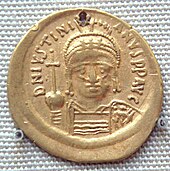 A photograph of a gold coin, showing a man wearing a crown holding up a cross with his right shoulder. On the top are letters in the Roman script.