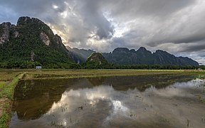 Reflet dans l'eau des montagnes de Vang Vieng, avec un ciel nuageux et des rayons crépusculaires illuminant le sommet d'une colline et une fine portion de rizières vertes, le soir, pendant la mousson.