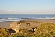 Photographie d'un bunker en ciment, à l'état de ruine, situé à l'abri d'une dune, au bord d'une plage.