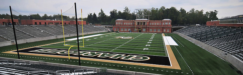 A wide photograph of an empty stadium. Stands are on the left, a brick building behind an American football playing surface is near the center, and home side stands with a press box are on the right.