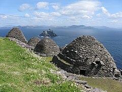 Photographie en couleurs de trois bâtiments en forme de dôme, appareillés de pierres grises et blanches, un pré s'étendant sur la gauche, une mer et des îles visibles sur la droite.