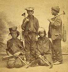 A studio portrait of four Afro-American climbing boys from New York, with brushes and scrapers, Two are standing and two are kneeling. They look between eleven and fourteen years old, wear rough clothes and battered hats and caps.