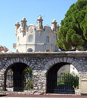 Vue d'une petite tour de château en fond avec au premier plan une muraille en pierre avec arcades barrées de fer forgé.