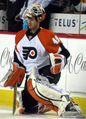 An ice hockey goal tender leans on one knee by the edge of an ice hockey rink. He is wearing a white uniform with orange shoulders and '49' on the sleeve, and a stylised 'P' on his chest, with a lot of padding visible.