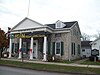 A light brown stone building with a pointed roof, green shutters and white columns on the front. In front of it are a sidewalk, a blue marker with gold writing, and a portion of street. Black letters above the front columns read "Holland Land Office Museum", which is also on a banner across the front.