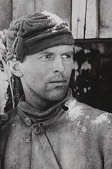 A head and upper torso portrait of Tryggve Gran, aged around 25, looking over his left shoulder with an intense expression, wearing winter clothing with snow on his woollen cap.