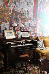 Farbfotografie des restaurierten James-Ensor-Hauses. Hinter einem Harmonium und einem Sofa ist an der Wand ein Ausschnitt des Gemäldes „Der Einzug Christi in Brüssel“ zu sehen.