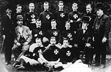 Fifteen men posing across three rows. Eleven of the men are wearing a football kit with a Maltese Cross on the breast. The other four are wearing suits and top hats