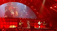 AC/DC with touring, lead vocalist Axl Rose, aged 44, shown in right profile both on screen and centre stage. He wears a black hat, T-shirt and jeans with gold jewellery. Chris Slade on drums is shown on screen but obscured on stage. Stevie is at right stage, Angus in white shirt and dark shorts to Rose's left, Williams in black at stage left.