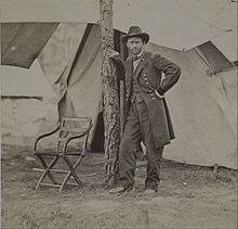 Photographie d'un homme en uniforme militaire prenant la pose en s'appuyant contre un arbre devant une large tente