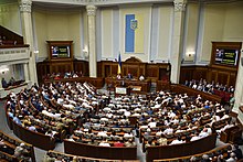 Vogelperspektive auf das vollbesetzte Parlament mit einem Redner am Pult. Die Wände bestehen aus hellem Marmor und haben Säulen. Auf beiden Seiten vom Hauptpult sind Bildschirme mit Übertragungen der Rede aufgebaut. An der linken Seite des Fotos erkennt man einen Rang.