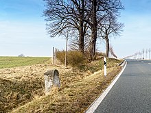 In offener Landschaft steht ein alter Kilometerstein und eine Gruppe älterer Bäume an einer modernen Landstraße. Die Bäume und ein kleiner Erdwall bildet eine kleine Bucht an der Straße.