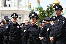 Im Vordergrund der Farbfotografie stehen drei Polizisten, jeweils ein Mann links und rechts vom Foto und eine Frau in der Mitte, mit Blick an der Kamera vorbei. Im Hintergrund sind weitere Polizisten und eine orthodoxe Kirche zu erkennen. Alle Polizisten tragen die schwarze Uniform mit einem runden Abzeichen an ihrer linken Brust.