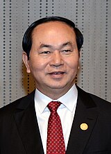 Trần Đại Quang looking a bit to the right, wearing a black suit, white shirt and a red tie while having a red-yellow pin on his right chest.