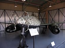 A twin engine jet aircraft pictured from front-left sitting on the ground in a hangar. The paint scheme of the aircraft is camouflage of various brown and green colours. Two antennas are protruding from the nose of the aircraft. The white number "305" is visible on the nose of the aircraft.