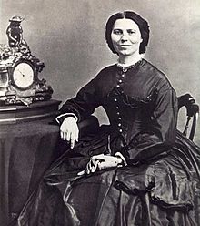 19th century photograph of a woman seated with her arm resting on a table. Her dark hair is neatly partedin the middle. She is smiling slightly