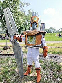 Photo d'un homme au visage peint en bleu dans un costume blanc et orange, muni d'un diapason.