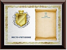 A commemorative plaque granted to Rescuer Cities in the shape of a horizontal rectangle. The base of the plaque is made of laminated composite material, on which a rectangular plate of white metal is placed. The right half of the plate depicts an unfolded scroll with the text of the Decree of the President of Ukraine on awarding an honorary award. In the left half of the plate, an honorary award without a block is depicted, below it is an inscription in two lines in blue letters: "______________SAVIOR CITY". On the edge of the plate there is an ornament in the form of golden laurel leaves.