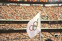 A white flag with five differently colored interlocking rings in the center is seen atop a flagpole, against the backdrop of a stadium filled with spectators.