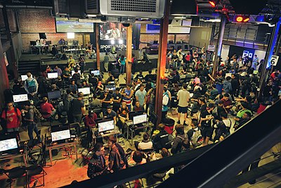 Overhead shot of a room full of people. In the foreground, several monitors are set up on tables and people are competing around each one. In the background, a light and camera setup focuses on a featured match, which is broadcast on a screen that audience members are watching.