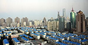 A cityscape seen from a height. In the foreground are low-rise buildings with blue roofs, giving way at the rear to higher-rising towers of different shapes and materials.