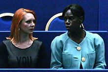 À gauche, Marina Anissina, à droite, Surya Bonaly portant une veste bleue. Les deux femmes sont assises devant une rangée de sièges vides.