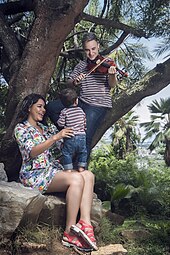 Photographie en extérieur d'une femme tenant un petit garçon sur ses genoux ; celui-ci porte son attention sur son père, installé sur une branche d'arbre en train de jouer du violon