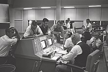 Photographie en noir et blanc de Kraft, assis à sa console, échangeant avec ses collègues. Gene Kranz, Elliot See et John Aaron sont notamment présents.