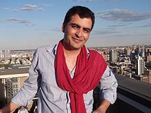 A photograph of Manav Kaul shot in the year 2010. He is wearing a light blue shirt and red scarf. In the background is a cityscape and a clear sky.