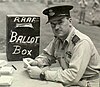 Man in military uniform with peaked cap seated at a desk beside a box marked "RAAF Ballot Box"