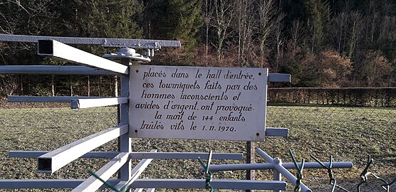 Plaque blanche apposée au tourniquet en métal et indiquant : « Placés dans le hall d’entrée, ces tourniquets faits par des hommes inconscients et avides d’argent ont provoqué la mort de 144 enfants brûlés vifs le 01.11.1970 »