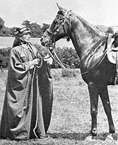 A black-and-white photograph of a European woman dressed in Bedouin robes and head covering, standing in front of a dark horse equipped with a bridle and saddle.