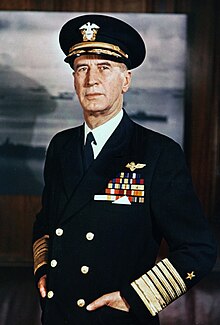 A middle aged man in naval uniform with tie, cap and awards standing with his hands in his pockets, in front of a large framed photograph of ships at sea. Color photograph