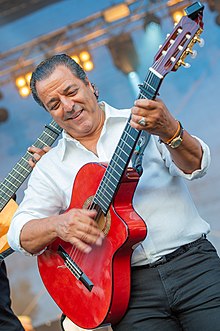 Jalloul Bouchikhi wearing a white shirt and dark pants, playing a red acoustic guitar onstage