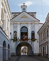 Frontale Farbfotografie eines blau-weißen Gebäudes mit einer Tordurchfahrt auf einer schmalen Straße. An beiden Straßenseiten stehen alte Gebäude. Über dem Tor ist eine Etage mit drei Rundbogenfenstern, in der Menschen stehen. Im darüber liegenden Giebelfeld ist ein goldenes Dreieck mit goldenen Strahlen und der lateinischen Aufschrift „Matea Misericordia“. Der hintere Dachbereich ist rechteckig und auf dem Giebel ist ein kleiner Glockenturm mit einem goldenen Jesuskreuz.