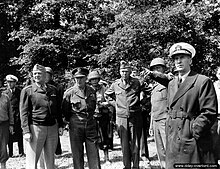 Black and white photograph, five men in varying military uniforms are in the foreground in front of some other men and a forest