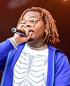 A man with brownish dreadlocks in a blue zip through sweat top and white shirt holding a microphone