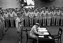 MacArthur is seated a small desk, writing. Two men in uniform stand behind him. A large crowd of men in uniform look on.