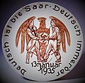 Gedenkteller zur Saarabstimmung: Brauner Reichsadler mit Hakenkreuzemblem, davor reichen sich ein NS-Parteigenosse (links) und ein Saarbergmann mit Grubenlampe (rechts) die Hand. Unterhalb ist das Datum der Abstimmung eingetragen: „13. Januar 1935“: Entlang des Randes befindet sich die Umschrift „Deutsch ist die Saar – Deutsch immerdar“ nach dem „Saarlied“ von Hanns Maria Lux. Waechtersbacher Steingut, 1935