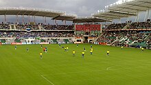 Farbfotografie in der Obersicht von einem Teil des Fußballstadions mit vollen Tribünen und einem Tor im linken Bereich. Zwischen den Tribünen in der Mitte ist ein großes Werbeplakat. Im Vordergrund spielen Menschen in gelben und blauen Trikots um einen Ball, der sich in der Bildmitte befindet. Der Schiedsrichter im roten Trikot steht auf dem Rasen weiter links.