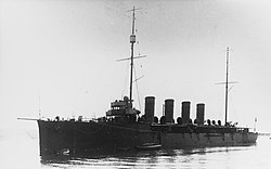 A cruiser sits still in the water with the coastline in the background. Two masts and four funnels sit prominently in the middle of the ship.