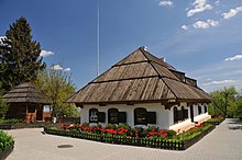 Farbaufnahme vom einstöckigen Gebäude mit seinem Holzdach und umzäunter Wiese, die rote Blumen hat. Im linken Hintergrund ist noch eine kleine Holzhütte.